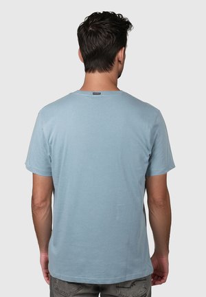 Mann med mørkt hår som bærer en lyseblå kortermet T-skjorte, sett bakfra, stående mot en ensfarget lys bakgrunn.