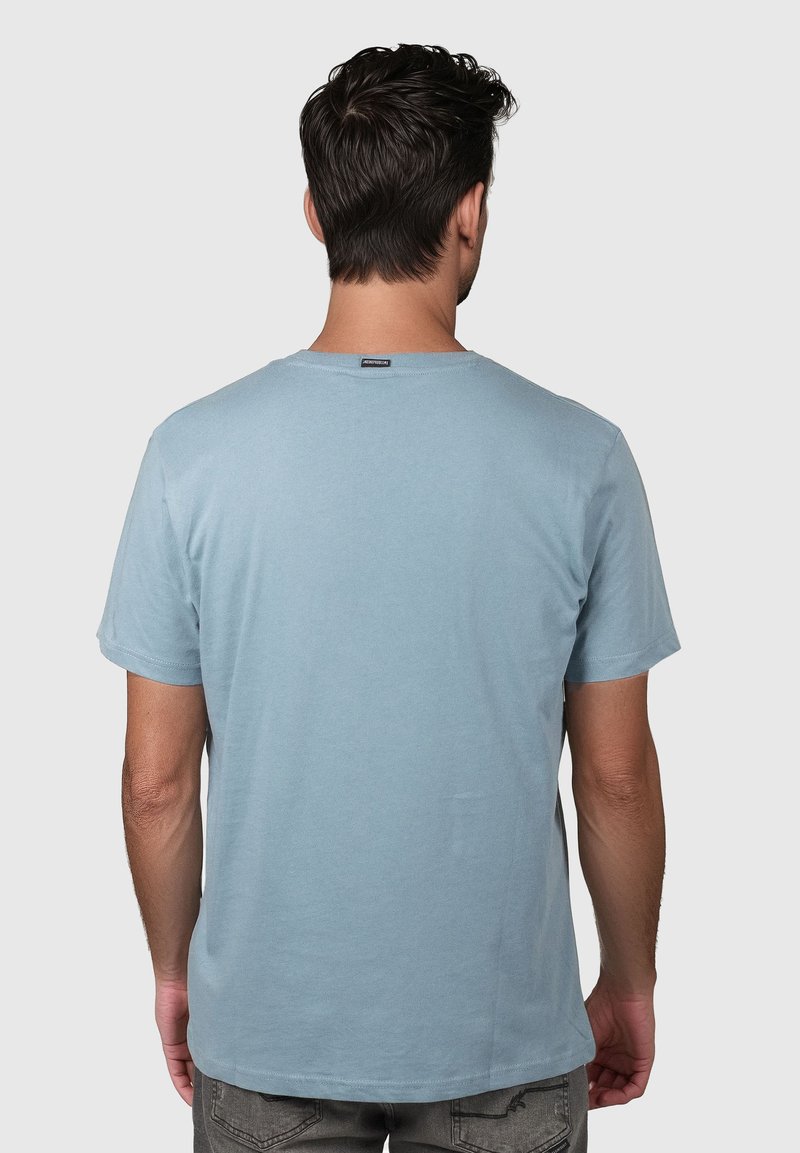 Muž s tmavými vlasy nosící světle modré tričko s krátkým rukávem, otočený zády, stojící na jednoduchém světlém pozadí.