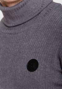 Szary sweter z golfem, o prążkowanej strukturze, z okrągłą czarną łatką z logo. Teksturowany materiał dzianinowy i dopasowany krój.