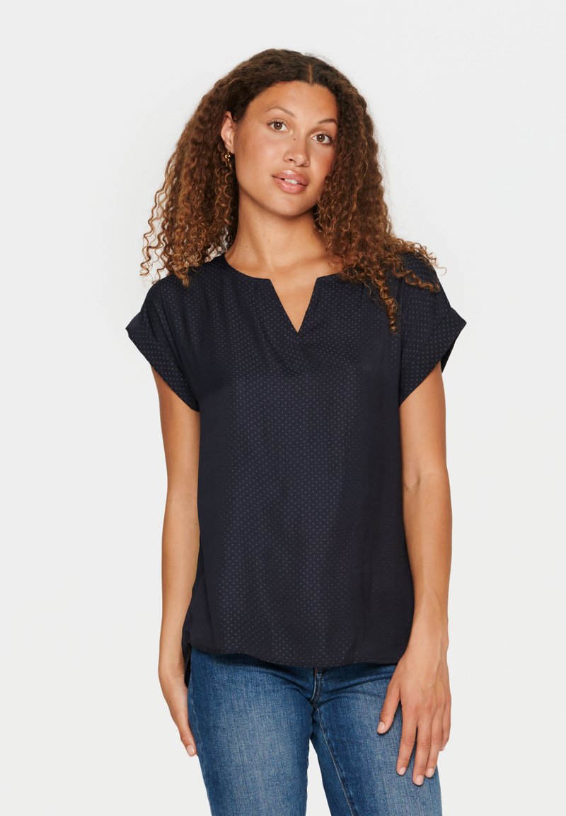Blusa navy con scollo a V e maniche corte, realizzata in tessuto leggero con un sottile motivo a pois. Vestibilità casual, perfetta per essere indossata a strati.