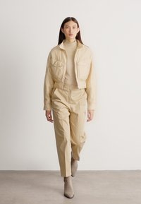 Γυναίκα που περπατά προς τα εμπρός, φορώντας μονοχρωμική μπεζ εμφάνιση με κοντό μπουφάν, παντελόνια ψηλής μέσης και μποτάκια μέχρι τον αστράγαλο, σε απλό φόντο.