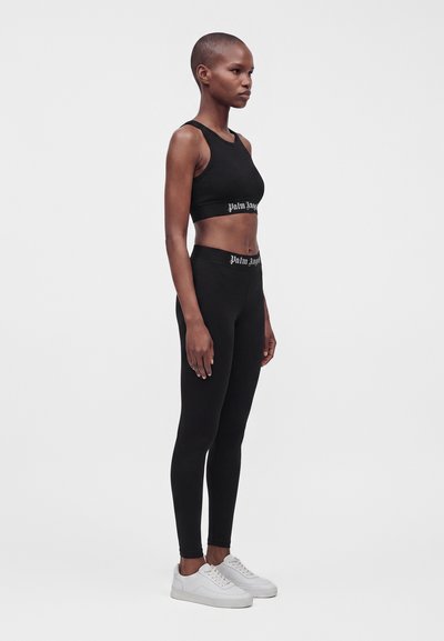Γυναίκα που φοράει μαύρο αμάνικο crop top και κολάν με λευκό κείμενο στις ζώνες μέσης, με λευκά αθλητικά παπούτσια, σε απλό ανοιχτό φόντο.