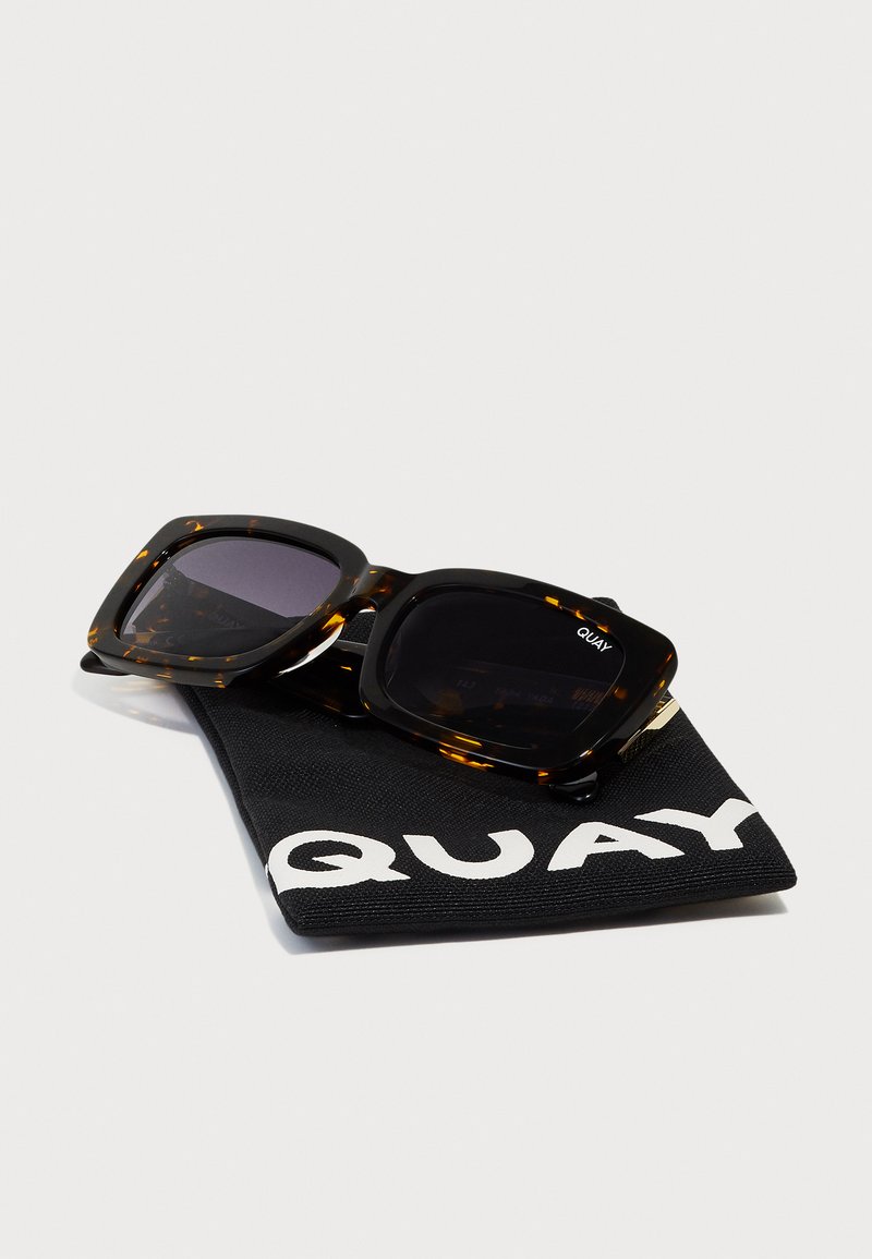 Schildpatt-Sonnenbrille mit dicken quadratischen Rahmen, dunklen Gläsern und einem schwarzen Tragetasche mit einem weißen Logo.