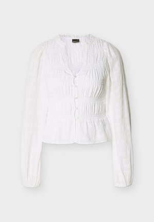 Λευκή μπλούζα με μακριά μανίκια, με σούρες στο ύφασμα, κουμπιά μπροστά, στρογγυλή λαιμόκοψη και ελαφρώς φουσκωτούς ώμους.