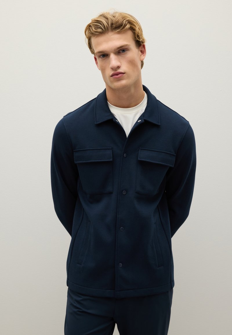 Giacca button-up blu navy in misto cotone, con due tasche sul petto, tasche laterali e colletto rigido. Tessuto morbido.