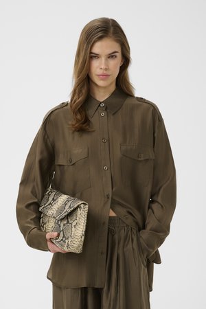 Frau mit langen braunen Haaren, die ein lockeres olivfarbenes Hemd mit Knöpfen und eine Hose trägt und eine Clutch mit Schlangenledermuster hält.