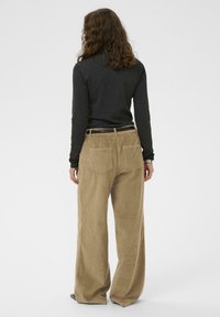 Persona con cabello castaño ondulado, vistiendo una blusa de manga larga oscura y pantalones anchos de pana beige, de pie de espaldas sobre un fondo blanco.