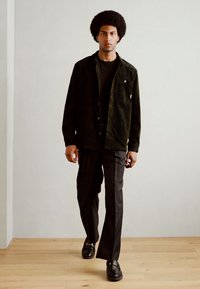 Camisa chaqueta de pana negra con cierre de botones, camiseta negra, pantalones negros y mocasines negros, sobre un fondo claro y neutro.