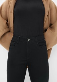 Jeans neri a vita alta con una texture liscia, caratterizzati da una chiusura anteriore con bottone, tasche laterali e dettagli di cucitura. Abbinati a un top nero.