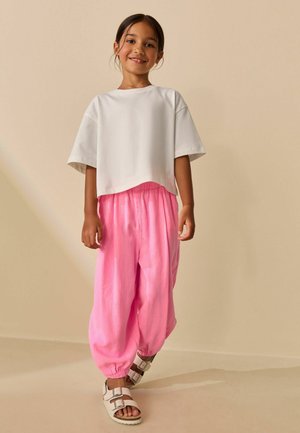 Usmívající se dívka stojící, oblečená v volném bílém krátkém topu, jasně růžových volných kalhotách a bílých sandálech s přezkami na béžovém pozadí.