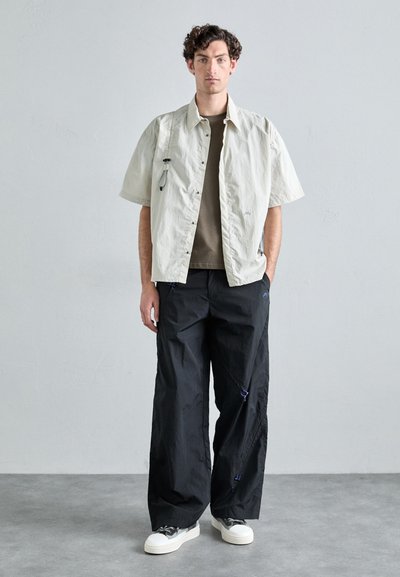 Chemise beige légère à manches courtes avec un t-shirt gris en dessous, accompagnée de pantalons noirs amples avec des accents bleus et des baskets blanches.