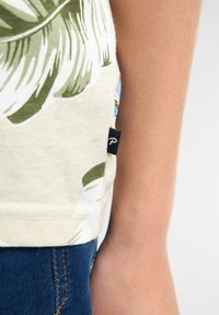 Kortærmet, lysebrun skjorte med grønne palmeblade. Vævet mærke i mørkeblå på sidesømmen. Kombineret med blå denimshorts.