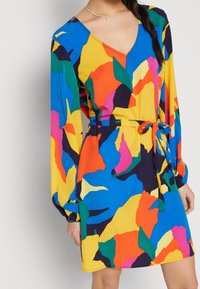 Robe multicolore avec un décolleté en V, des manches longues, une taille cintrée avec un lien, et un motif abstrait audacieux en bleu, jaune, rouge et vert.