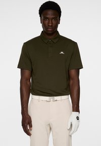 Ciemnozielona koszulka polo z kołnierzykiem i krótkimi rękawami, z białym logo na piersi. Nosi ją w połączeniu z beżowymi spodniami i białą rękawiczką golfową.
