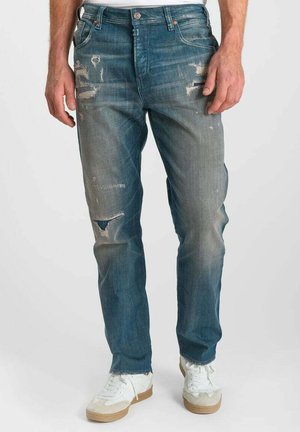 Man draagt vaalblauwe versleten spijkerbroek en witte sneakers, staand tegen een effen lichte achtergrond.