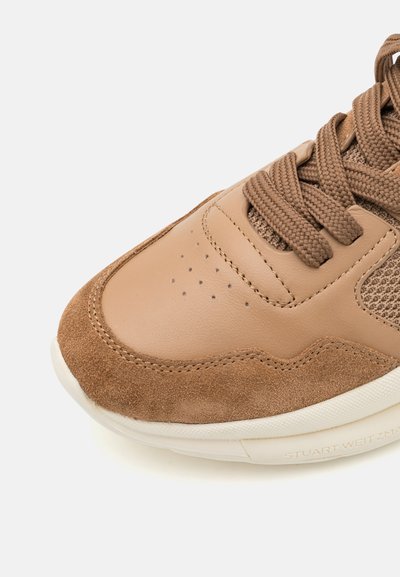 Sneaker con una toma in pelle beige, con accenti in suede, rete testurizzata e lacci tondi. La suola è bianca con dettagli di branding discreti.