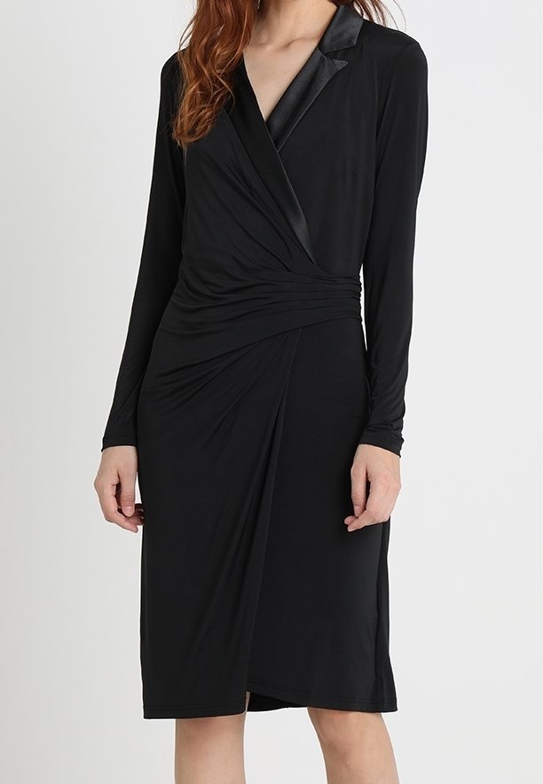 Femme portant une robe portefeuille noire à manches longues avec col en satin et détail froncé à la taille, longueur genou, sur fond uni.