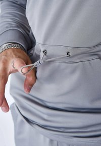 Felpa sportiva grigio chiaro con chiusura a coulisse. Presenta occhielli in metallo e una texture del tessuto elegante, con una mano di una persona che regola la coulisse.