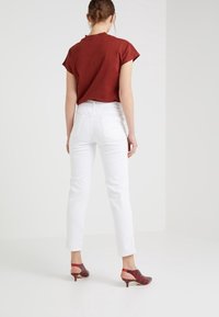Mujer de pie, de espaldas, con una camiseta corta color óxido, jeans blancos de tiro alto y zapatos de tacón bajo tipo slingback rojos.