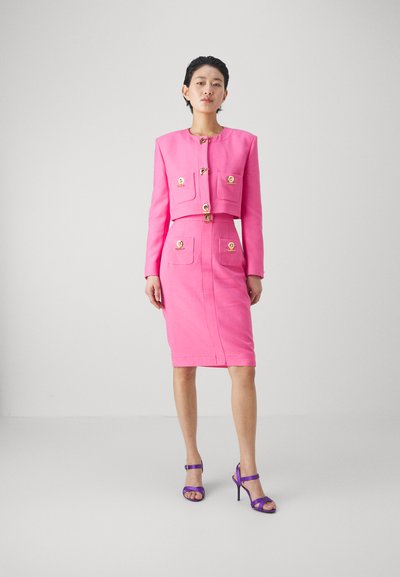 Costume rose sur mesure avec veste courte, boutons dorés et poches. Design de jupe crayon, associé à des sandales à talons hauts strappées violettes.