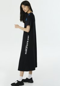 Zwart, oversized T-shirt jurk met korte mouwen, voorzien van witte tekst langs de zijkant en blauwe accenten op de schouders. Gladde stof.