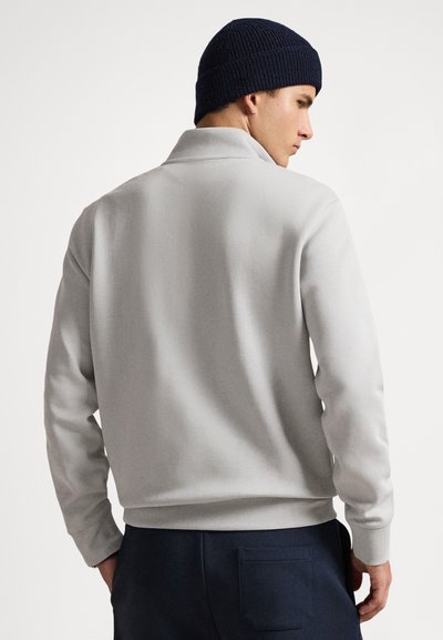 Άνδρας με ανοιχτόγκρι μακρυμάνικο πουλόβερ και σκούρο μπλε σκουφί, γυρισμένος ώστε να φαίνεται η πλάτη και το προφίλ του, μπροστά σε απλό φόντο.