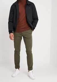 Mężczyzna w czarnej kurtce, brązowym swetrze z golfem, oliwkowych spodniach z podwiniętymi nogawkami i białych sneakersach, stoi na tle jednolitego jasnego tła.