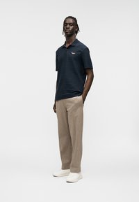Σκούρο ναυτικό μπλε κοντομάνικο πόλο μπλουζάκι με υφασμένες ρίγες και λεπτομέρεια λογότυπου, συνδυασμένο με ανοιχτό καφέ παντελόνια και λευκά αθλητικά παπούτσια.