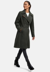 Dark green wool blend coat with a double-breasted front, large lapels, side pockets, and wood accent on collar. Worn over black jeans and ankle boots.