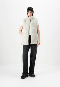 Vrouw staat met een mouwloze lichtgrijze nepbont gilet, een wit shirt met korte mouwen, zwarte wijde broek en zwarte chunky schoenen op een witte achtergrond.