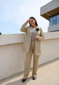 Bežový blazer s kapsami, doplněný o odpovídající kalhoty, nošený přes pruhované tričko. Obsahuje černý květinový akcent. Černé baleríny dotvářejí outfit.