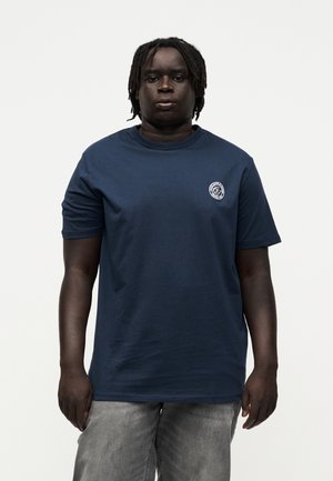 Sort person med korte dreadlocks bærer en mørkeblå t-shirt med et lille hvidt logo på venstre bryst og grå bukser, stående foran hvid baggrund.