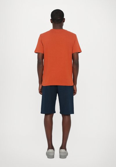 Rust-colored cotton t-shirt with short sleeves, paired with navy shorts. Model stands with back to the viewer, featuring gray sneakers.