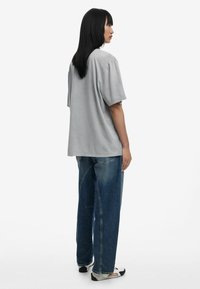 Sivá oversized tričko s krátkymi rukávmi, skombinované s voľnými modrými džínsami. Model stojí bokom, ukazujúc jednoduchý, ležérny siluet.