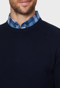 Marineblauer gerippter Pullover mit rundem Ausschnitt, über einem karrierten Hemd in Blau und Hellblau mit Button-Down-Kragen.