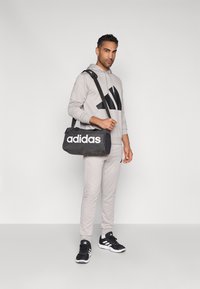 Μαύρη τσάντα Adidas τύπου duffel με λευκό λογότυπο, φορεμένη με γκρι φούτερ και παντελόνια τζόγκινγκ, σε ουδέτερο φόντο, που αναδεικνύει τον αθλητικό σχεδιασμό.