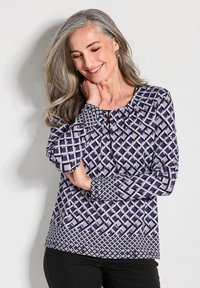 Langarmbluse mit lila geometrischem Muster, runder Halsauschnitt mit Schlitzdetails und strukturiertem Stoff. Kombiniert mit schwarzen Hosen.