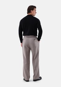 Schwarzer Pullover, hellgraue, maßgeschneiderte Hose mit glatter Textur und Rückenschlitzen. Das Modell steht mit den Händen in den Taschen und trägt schwarze Schuhe.