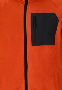 Oranje zip-up jack met een zwarte vierkante borstzak met ritsluiting, gemaakt van een gestructureerde stof. Verticale zwarte rits loopt over de voorkant.