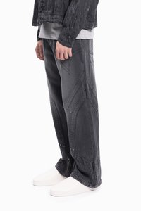 Szerokie nogawki, czarne dżinsy z ostrokątnymi wzorami przeszyć i detalami na zatrzask, zestawione z białymi butami i szarym koszulą.