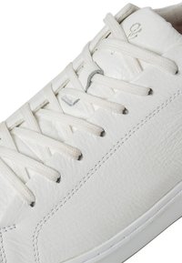 White leather sneaker with textured surface, round toe, and flat white laces. Features subtle stitching and minimal silver detailing.