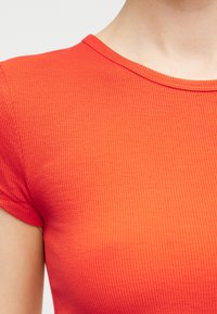 Κοντινό πλάνο ενός ατόμου που φοράει κοντομάνικη, ριγέ, φωτεινά πορτοκαλί μπλούζα με στρογγυλή λαιμόκοψη, μπροστά σε μια απλή φόντο.