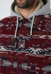 Bordeauxmønstret jakke med grå fleeceforet hætte, sorte knapper og geometriske designs i røde, blå og creme nuancer.