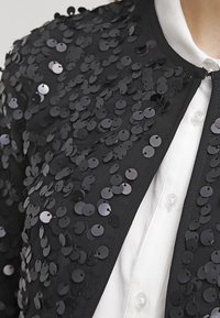 Černá bunda zdobená kruhovými flitry, s kulatým výstřihem, otevřeným předním dílem a bílou košilí pod ní.