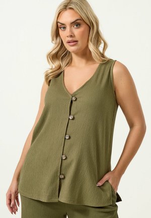 Frau mit blondem, welligem Haar, die ein ärmelloses, olivgrünes, knöpfbares Oberteil und passende Hosen trägt und mit einer Hand in der Tasche posiert.