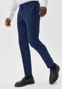 Jūrų laivyno spalvos kelnių slim-fit dizainas, su lygiu paviršiumi ir priekinių kišenių, derinamos su juodais loafer'iais ir tamsiomis kojinėmis.