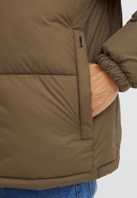 Smeđi pufasta jakna s glatkom teksturom, koja ima bočni džep, elastične manžete i blago podloženi dizajn.