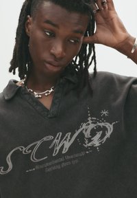 Black polo shirt with a collar, featuring a logo printed in gray. The fabric appears textured, with a relaxed fit and visible stitching details.