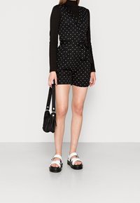 Svart prickig romper med v-ringning och bälte i midjan. Bärs över en svart långärmad tröja. Matchad med vita sandaler och en liten svart väska.