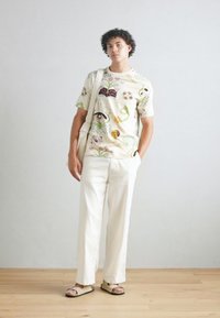 White floral-patterned t-shirt, cream-colored linen pants, beige sandals; model stands with a shoulder bag against a neutral background.